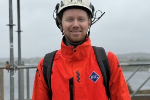 En mann står på en byggeplass og smiler mot kameraet. Han har på seg hvit vernehjelm, en oransje arbeidsjakke med refleks og en svart ryggsekk. I bakgrunnen er det stillaser og overskyet vær.