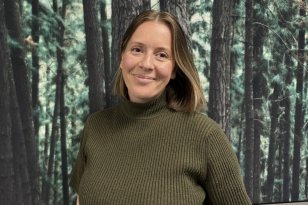 En kvinne med kort, lyst brunt hår står foran et stort bilde av en tett furuskog. Hun smiler svakt og ser rett mot kamera. Hun har på seg en olivengrønn, ribbestrikket topp med høy hals og korte ermer.