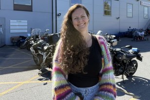 En kvinne med langt, brunt, krøllete hår står utendørs foran en bygning med motorsykler parkert utenfor. Hun smiler mot kamera. Hun har på seg svarte klær og en fargerik, stripete cardigan. Det er solskinn og blå himmel.