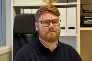 En mann med briller og rødt hår og skjegg sitter i en kontorstol på et kontor. Han har på seg en mørk strikkegenser og ser rett mot kamera. Bak ham står hyller med permer, esker og kontorutstyr.