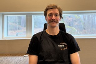 En mann sitter ved siden av et skrivebord og ser rett mot kamera. Han har bart og kort, brunt hår og har på seg en svart t-skjorte med logoen «Slevik Trevare». Bak ham er et vindu med utsikt mot skog.