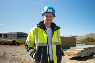 En mann står med hendene i lommene og smiler til kamera. Han har på seg hjelm og refleksjakke, og står på et stort åpent område med byggematerialer i bakgrunnen.
