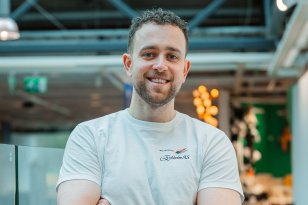 En mann i hvitt arbeidstøy står med armene i kors og smiler til kamera.