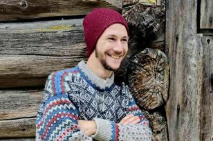 Mann med tradisjonell ullgsenser og rød lue lener seg mot en vegg av tømmer