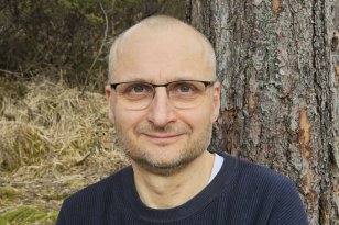 Mann med briller og svart genser står foran et tre. I bakgrunnen er det skog.