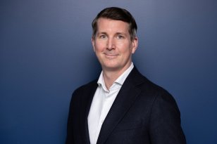 Mann med hvit skjorte og svart dressjakke står foran en blå vegg