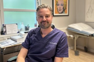 En mann i mørkeblå legeuniform sitter i et lyst legekontor og smiler rolig mot kamera. Bak ham sees en undersøkelsesbenk, medisinsk utstyr og et fargerikt portrett av Marilyn Monroe på veggen.