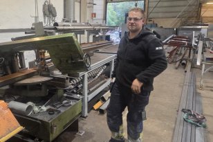 En mann står inne i et verksted omgitt av metallplater, rør og store maskiner. Han har på seg vernebriller, svart arbeidsjakke og mørke arbeidsbukser med refleksdetaljer. I bakgrunnen er det åpne porter som slipper inn dagslys og viser trær utenfor. Verkstedet er fullt av industrielt utstyr, og gulvet er støvete etter arbeid.