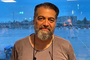 Mann med mørkt hår og skjegg står foran et vindu. Han smiler til kameraet.