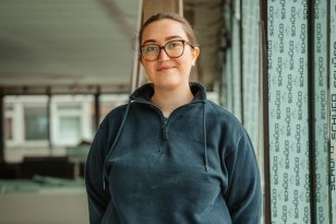 En kvinne med brunt, oppsatt hår og briller står med hendene bak ryggen og smiler til kamera. Hun har på seg arbeidsklær og står inne på en byggeplass.