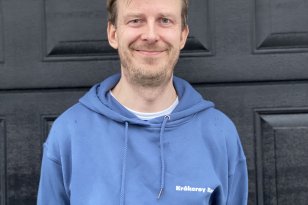 Rørlegger  Mann med blå hettegenser står foran en svart dør og smiler.