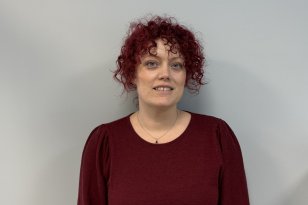 En kvinne med kort, krøllete rødt hår står foran en lys, nøytral vegg og ser rett mot kamera. Hun har på seg en mørk rød genser med lange ermer og et enkelt smykke rundt halsen. Ansiktsuttrykket er rolig og vennlig med et svakt smil.