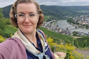 Lyshåret dame med briller står foran en landsby i bakgrunnen som ligger på hver side av en elv. Det er grønt og frodig i landskapet.