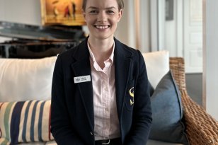 En ung kvinne i mørkeblå uniform sitter i en sofa. Hun har lyserosa skjorte på seg. Hun smiler mot kameraet.