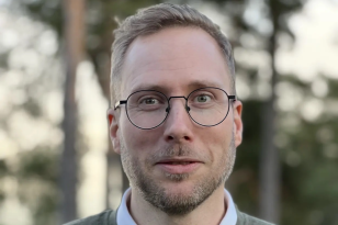 En mann står utendørs og ser rett mot kamera. Han har kort, lyst hår, kort skjegg og runde briller. Han har på seg en grå genser over en hvit skjorte. Bakgrunnen er litt uskarp med trær i dempet lys.