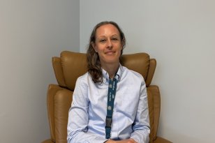 En kvinne i lyse klær med brunt, halvlangt hår sitter i en brun lenestol på et kontor eller møterom. Hun ser inn i kameraet og smiler svakt. Veggene er lyse.