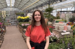 En ung kvinne med langt, mørkebrunt hår som henger løst over skuldrene, står i et drivhus omgitt av blomster og planter. Hun har på seg en rød T-skjorte og en svart genser knyttet rundt livet, og smiler mot kameraet.