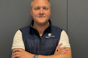 En mann står med armene i kors foran en mørkegrå vegg og ser mot kameraet. Han har på seg en hvit T-skjorte og en mørkeblå vest med Mopedbil Norge-logo. Han har klokke på håndleddet og tatoveringer på armene.