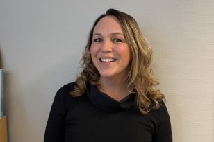 En kvinne med skulderlangt, bølgete hår står foran en lys vegg og smiler mot kamera. Hun har på seg en svart topp med høy hals og lange ermer.