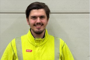 Mann med brunt hår og gul arbeidsjakke med refleks står foran en lys vegg og smiler til kamera