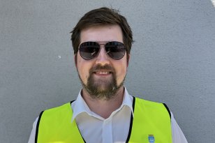 En mann med brunt hår og brunt skjegg smiler. Han har på seg solbriller med mørke glass, en gul refleksvest og en hvit t-skjorte med krage under. Han står inntil en grå vegg.
