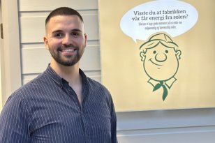 En mann med kort mørkt hår og skjegg står og smiler foran en lys vegg. Han har på seg en mørkeblå skjorte med hvite, tynne striper. Bak ham henger et skilt med en tegnet figur av en smilende mann og teksten: «Visste du at fabrikken vår får energi fra solen? Slik kan vi lage gode matvarer på en enda mer miljøvennlig og bærekraftig måte.»