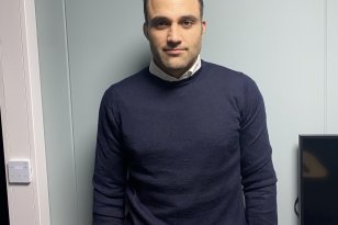 Mann med svart genser og mørkt hår står foran en kontorvegg