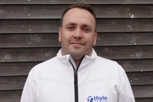 Mann som smiler til kamera med hvit arbeidsjakke med blå logo.