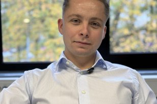 En ung mann med kort, lyst hår og lyseblå, stripete skjorte sitter foran et stort vindu og ser mot kameraet. Utenfor vinduet skimtes trær med høstfarger.