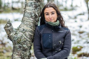 Damed med svart hår og svart fleecejakke og strikkevotter lener seg mot et tre i en skog med litt snø på bakken