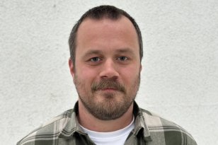 En mann står foran en lys murvegg og ser rett mot kameraet. Han har kort hår, skjegg og bart, og smiler svakt. Han har på seg en grønn- og hvitrutet flanellskjorte med en hvit T-skjorte under.