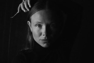 Skomaker Irene Dorthea Johansen Det er et svart-hvitt portrett av en ung kvinne med langt hår som faller bak skuldrene. Hun har et alvorlig uttrykk og ser rett mot kamera. Det ene armen er løftet, og hånden holder en liten metallgjenstand over hodet. Ansiktet er delvis opplyst fra venstre side, mens bakgrunnen og den andre siden av ansiktet er mørk. Hun har på seg en mørk, høyhalset genser.