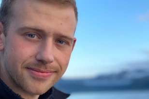 Bildet er et nært portrett av en ung mann som smiler svakt mot kameraet. Han har kort, lyst hår, blå øyne og lett skjeggvekst. Han har på seg en mørk jakke. Bak ham ser man havet og et blålig landskap i myk uskarp bakgrunn, trolig fjell i det fjerne. Himmelen er klar og blå.