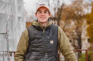 En mann med arbeidsklær og caps står og lener seg på en maskin. I bakgrunnen er det tydelig høst og overskyet. Mannen smiler til kamera.