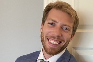 En mann med blondt hår og skjegg smiler bredt. Han har på seg dress - grå dressjakke, hvit skjorte, grønt slips og 17.mai rød sløyfe. Bak han er det en grå vegg og hvit dør.