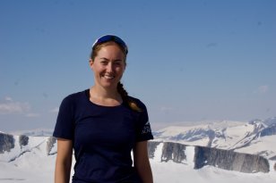 Dame med svart t-skjorte og solbriller står på et fjell, det er blå himmel og snø på fjellene rundt