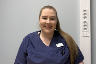 En kvinne med langt, brunt hår samlet i en høy hestehale sitter foran en lys grå vegg. Hun har på seg mørkeblå arbeidsuniform og smiler mot kameraet.