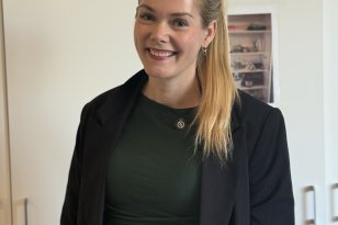 En smilende kvinne med langt, blondt hår i hestehale står foran et hvitt skap. Hun har på seg en mørkegrønn overdel og en svart blazer.