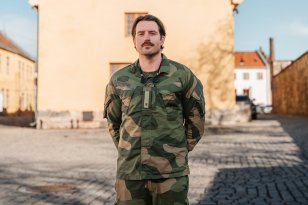 En mann med brunt hår og uniform står med hendene bak ryggen og smiler til kamera. I bakgrunnen er en lys, gammel bygning og blå himmel.