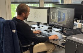 En mann i mørk genser sitter ved dataskjerm og jobber med tredimensjonale modeller. Han er på et kontor.