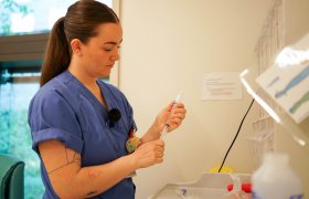 Ung kvinne med langt brunt hår i en hestehale og mørkeblå sykepleieruniform, står og trekker medisin inn i en sprøyte.