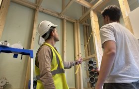 To personer står i et rom hvor reisverket i tre vises. Den ene personen har på seg gul refleksvest og hvit vernehjelm og peker en tommestokk mot den ene planken. Den andre personen har på seg hvit t-skjorte. Bilde er tatt nedenfra. De ser bort fra kamera.