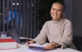 En ung kvinner med lyst hår i hestehaler sitter ved et bord og smiler, hun har et ark i hendene og Norske lover på pulten