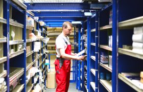 En mann i arbeidsklær står mellom to reoler fulle av reservedeler