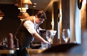 Mann i hvit skjorte og vest dekker et bord. Han er på en restaurant.