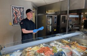 En mann i svart kokkeuniform og caps tar på seg et par blå plasthansker. Han står bak en disk fyllt med fisk og sjøkmat. I bakgrunnen er det et storkjøkken. 