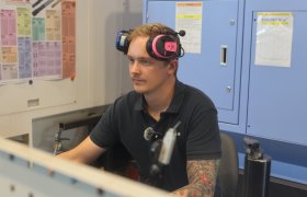 En mann sitter ved en dataskjerm i et industriverksted. Han har et rosa og svart hørselsvern på hodet og bruker en mørk arbeids­pique. I bakgrunnen står store CNC-maskiner og oppslag med tekniske tabeller. Personen jobber konsentrert foran skjermen.