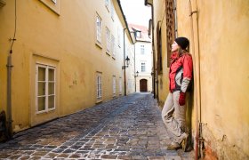 En ung jente ikledd rød jakke, lue og votter, lener seg mot en vegg. Bildet er tatt utendørs.