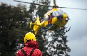 En person i gul hjelm og rød jakke ses bakfra. Personen ser på et helikopter.