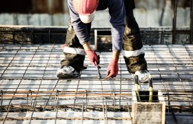 En person iført rød hjelm, hansker og øvrig arbeidstøy bøyer seg ned og jobber med armeringsjern på en byggeplass.